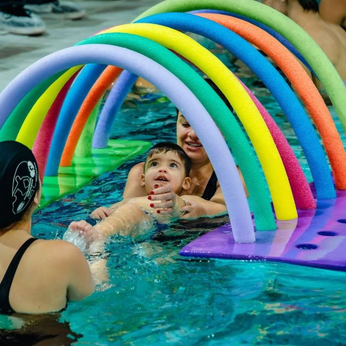 Criança nadando em piscina com apoio de duas mulheres, passando por túnel colorido feito com espaguetes de piscina durante aula de natação infantil.
