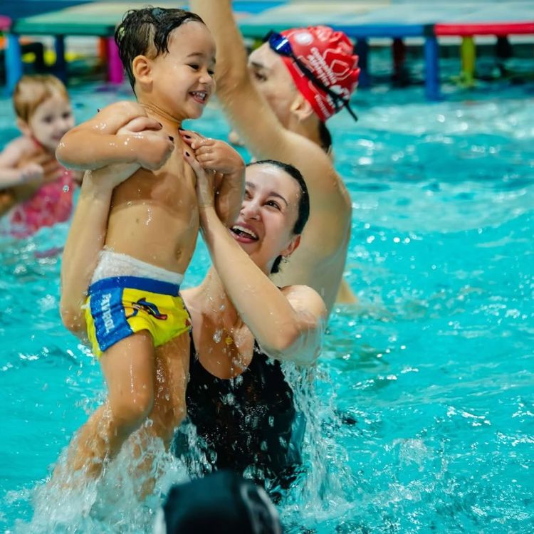 Mulher levantando uma criança sorridente dentro da piscina durante aula de natação infantil, com outros participantes ao fundo.