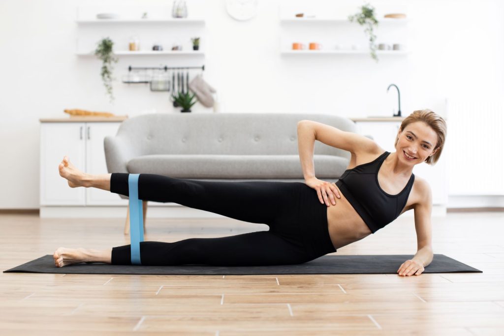 Mulher deitada de lado em um tapete realiza exercício com faixa elástica para fortalecimento do glúteo médio.  Essa prática é indicada para corrigir desequilíbrios musculares que contribuem para o joelho valgo