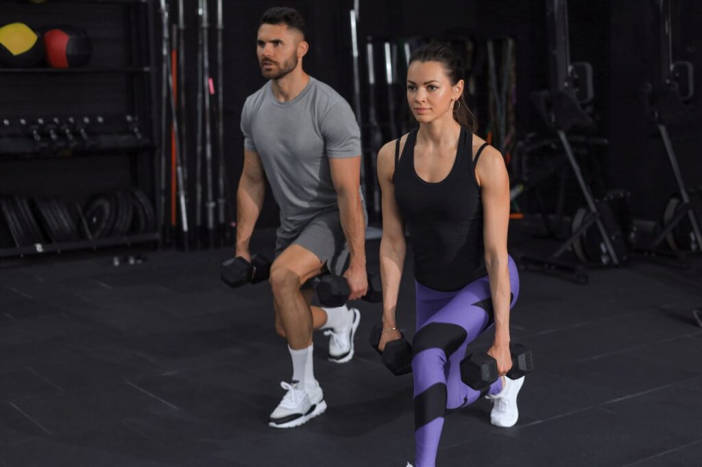 Homem e mulher fazendo agachamento com halteres em academia, vestindo roupas de treino cinza e roxa, com equipamentos ao fundo, destacando treino para perda de gordura e fortalecimento muscular.