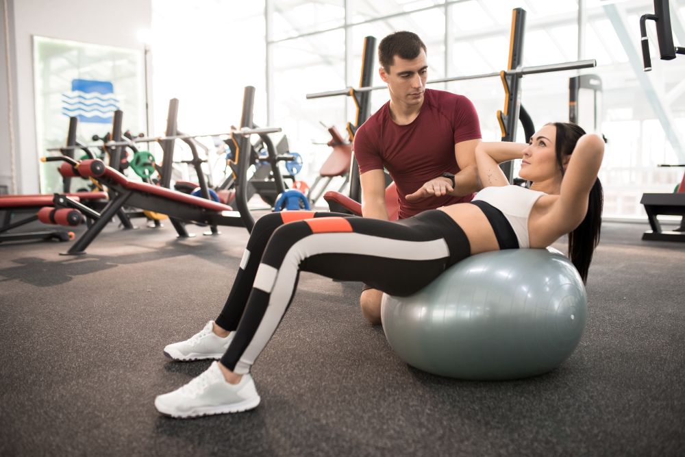 Mulher fazendo abdominal com bola de Pilates em academia, com treinador ajustando postura, vestindo top branco e legging preta, com equipamentos ao fundo, destacando treino para perda de gordura e fortalecimento do core.