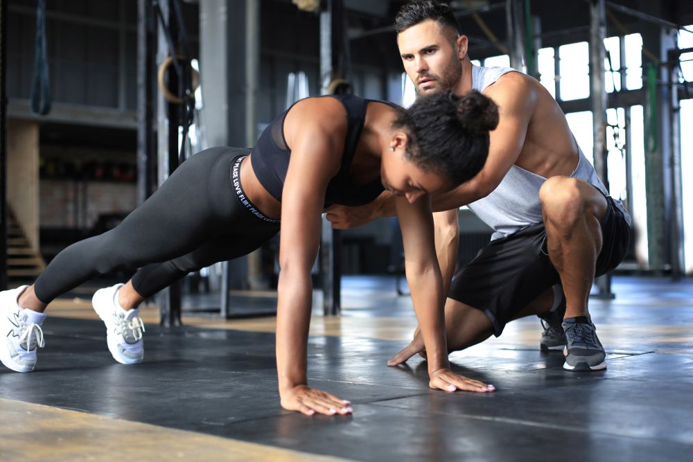 Mulher fazendo flexão em academia com treinador corrigindo postura, vestindo roupa de treino preta e tênis brancos, em ambiente com equipamentos e luz natural, destacando treino para perda de gordura corporal e fortalecimento.