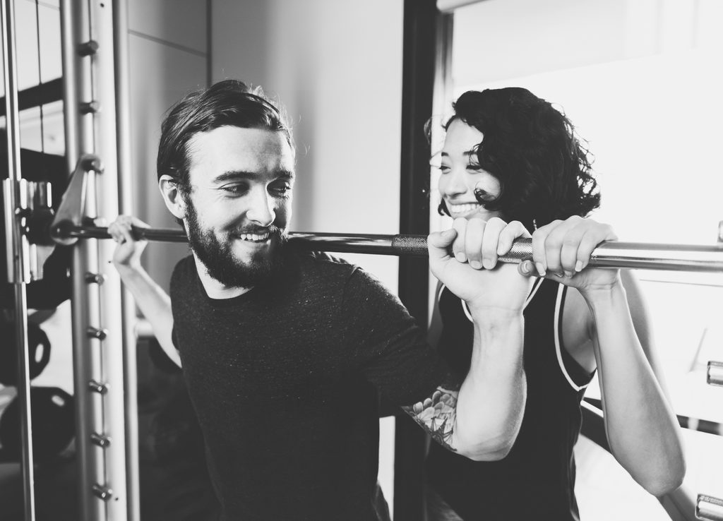 Homem realiza exercício com barra enquanto mulher auxilia e sorri em academia.