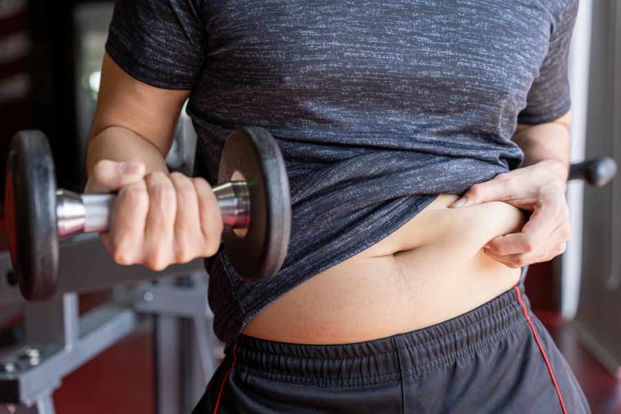 Pessoa em uma academia faz exercício de bíceps com um halter enquanto puxa a camisa para mostrar a gordura abdominal. A cena destaca o esforço para melhorar a forma física.