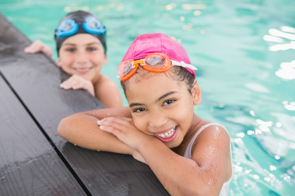 Crianças sorrindo na borda da piscina aquecida com toucas coloridas e óculos de natação durante aula de natação infantil, representando atividades infantis em Ribeirão Preto na Cia Athletica.