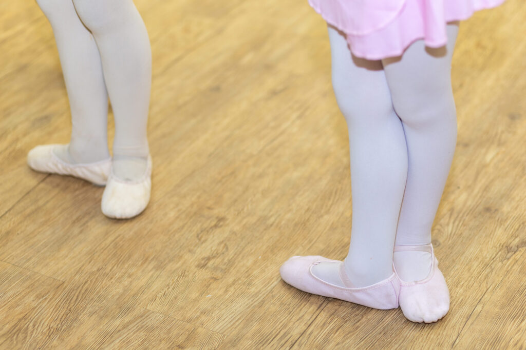 Pés de crianças com sapatilhas de ballet em posição básica sobre piso de madeira durante aula de dança.