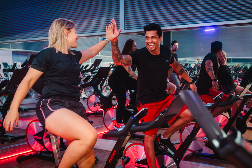 Pessoas praticando ciclismo indoor em academia moderna, sorrindo e trocando um high five durante aula coletiva de spinning, com iluminação vermelha e espelhos ao fundo, espaço amplo e iluminado, considerada uma das melhores academias de Ribeirão Preto.