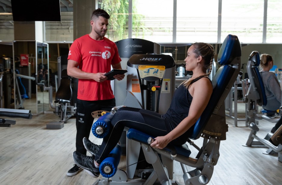 Personal trainer da Companhia Athletica em Ribeirão Preto orientando aluna durante treino personalizado em aparelho de musculação, com tablet em mãos e ambiente moderno ao fundo.
