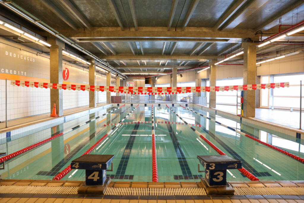 Benefícios da natação para crianças, adultos e idosos. Piscina coberta vazia, com raias delimitadas por divisórias vermelhas, brancas e pretas, blocos de partida numerados e iluminação natural pelas janelas laterais.