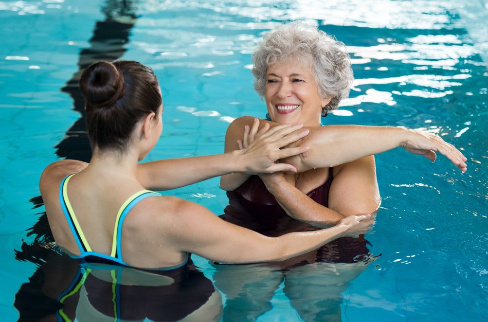 Instrutora auxilia idosa durante exercício de hidroginástica em piscina.