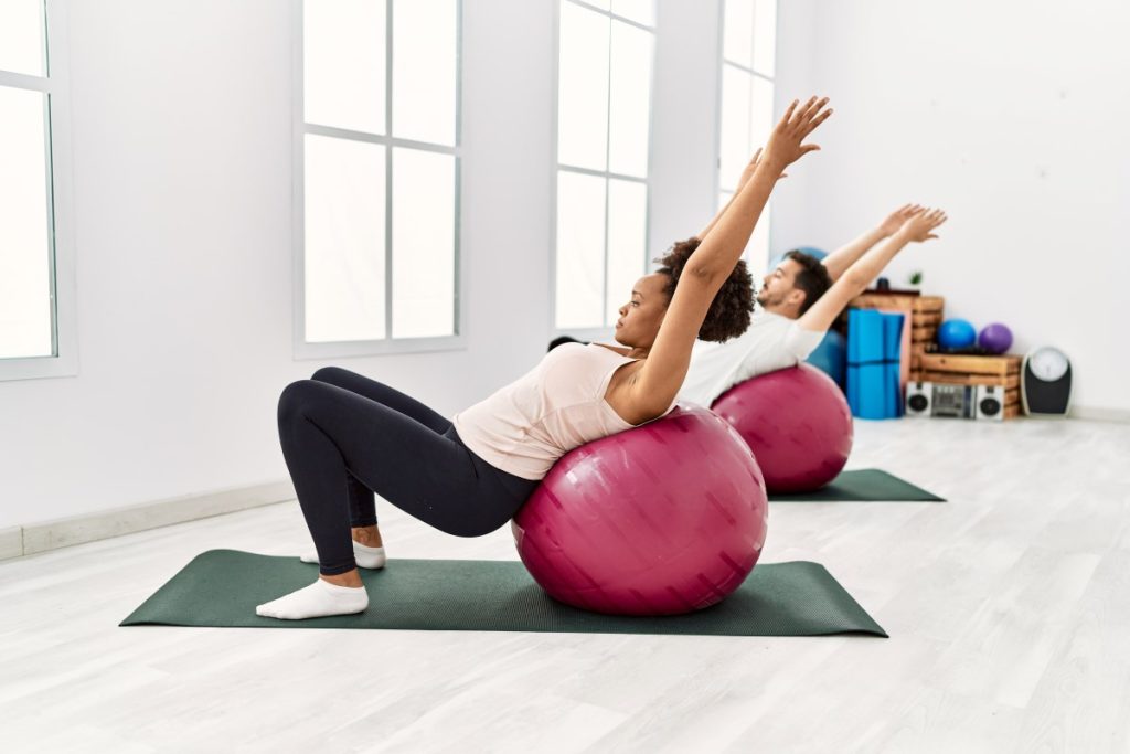 uma mulher e um homem, realizando exercícios de equilíbrio utilizando bolas de pilates em uma sala de exercícios.