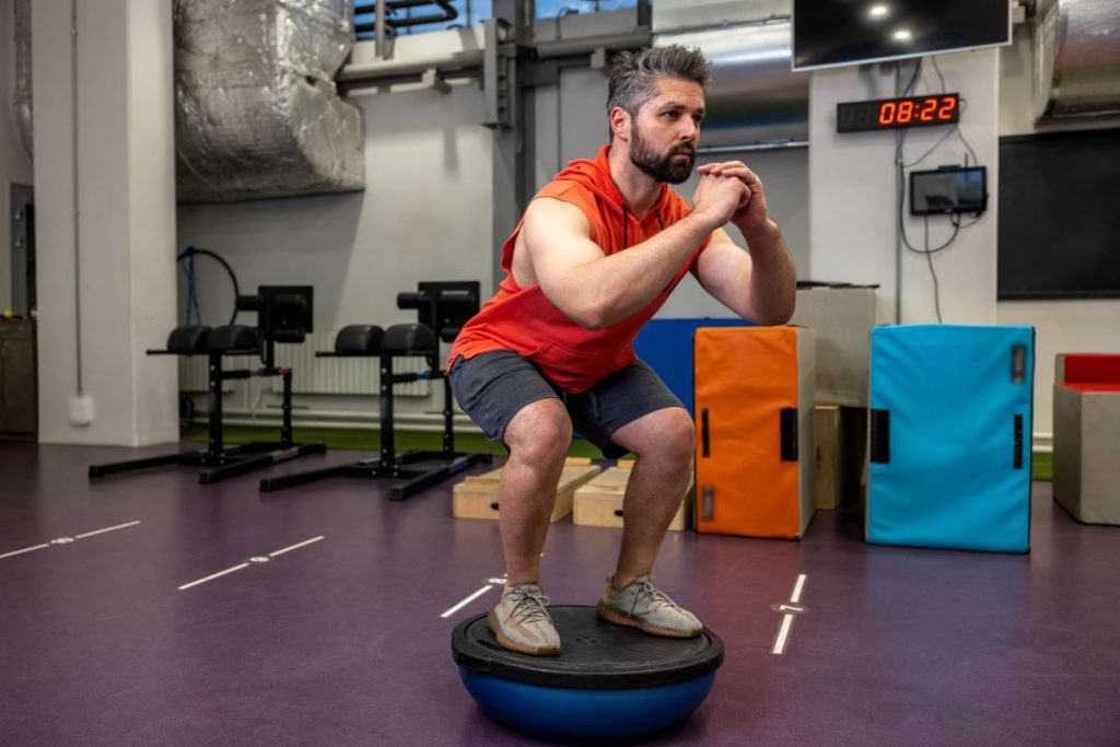 um homem realizando exercícios de equilíbrio usando um BOSU, uma plataforma com base semiesférica que desafia a estabilidade.