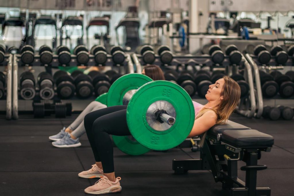 uma mulher está realizando o exercício de elevação pélvica com barra (também conhecido como hip push) em uma academia.