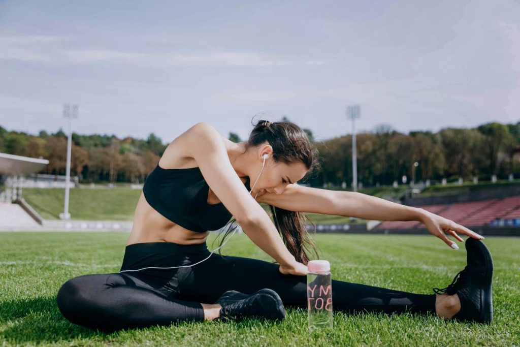 mulher está realizando um exercício de alongamento ao ar livre em um campo de grama, enquanto usa fones de ouvido