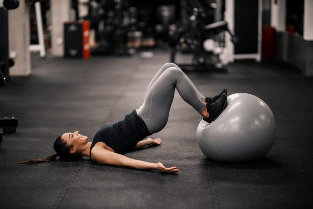 mulher deitada de costas em um chão de academia, realizando um exercício de elevação pélvica com o auxílio de uma bola de pilates