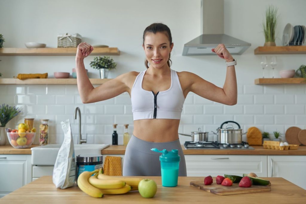 mulher com roupas de academia.  À sua frente, uma bancada exibe alimentos saudáveis, incluindo banana, maçã verde, morangos, pepino e um shake, fornecendo opções ideais do que comer antes do treino.