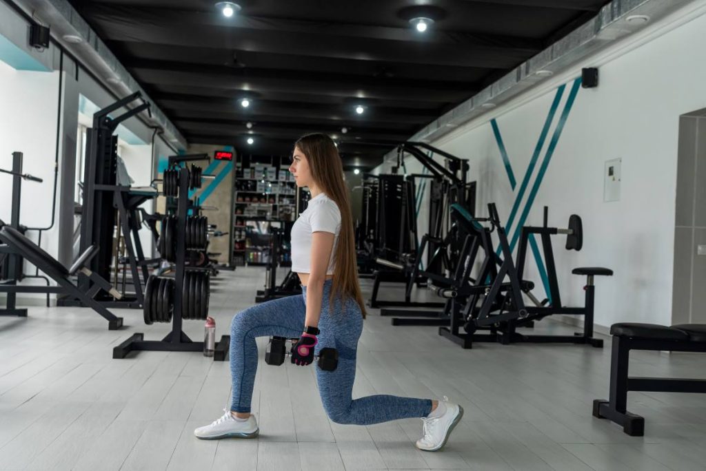 mulher realizando um exercício de afundo com halteres, com ênfase na musculatura posterior da coxa.