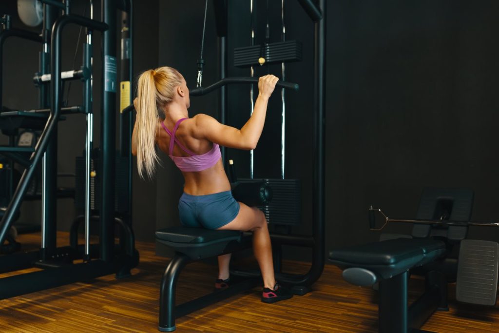 mulher loira, usando top roxo e shorts azul, realizando o exercício de puxada frontal em uma máquina de academia.