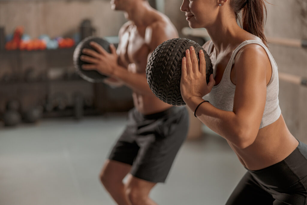 Funcional emagrece? Descubra como o treino funcional ajuda na perda de peso Homem e mulher realizam exercício funcional em academia, segurando uma bola de treino próxima ao peito enquanto executam movimento de agachamento.