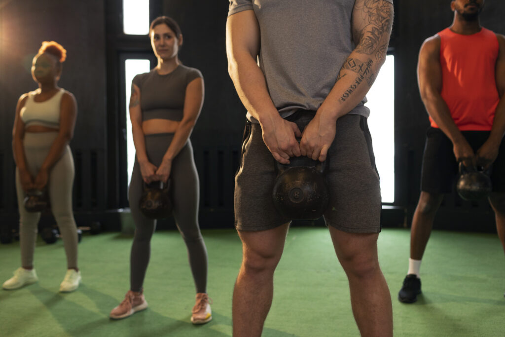 Grupo de alunos realiza exercício com kettlebell durante aula coletiva de treino funcional em academia.