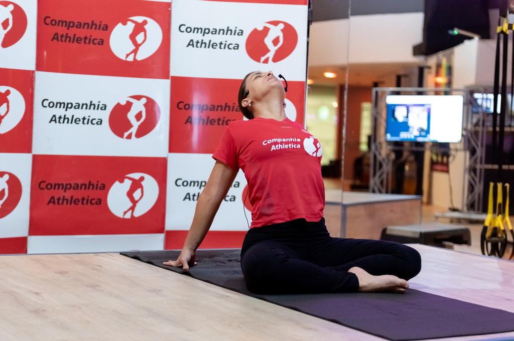 Professora da Cia Athletica realizando postura de yoga sentada com extensão de coluna sobre colchonete, vestindo camiseta vermelha da Companhia Athletica, em ambiente da academia em Ribeirão Preto.