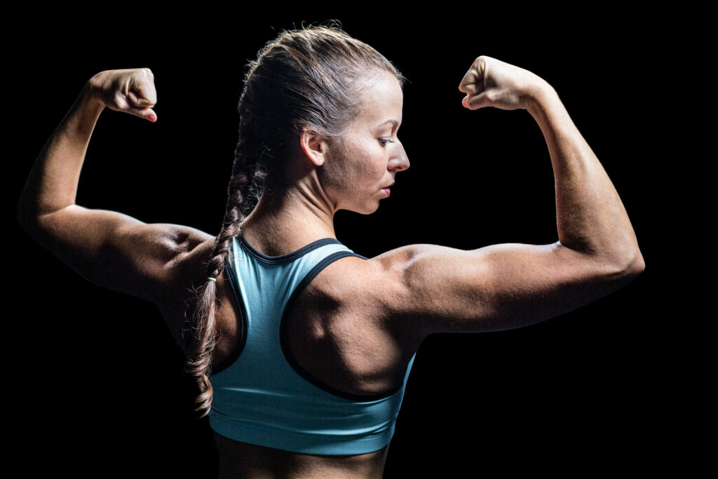 Exercícios para braços: o ajuste simples que pode acelerar sua hipertrofia Mulher atleta flexiona os braços e mostra os músculos das costas e bíceps contra fundo preto durante pose de treino.