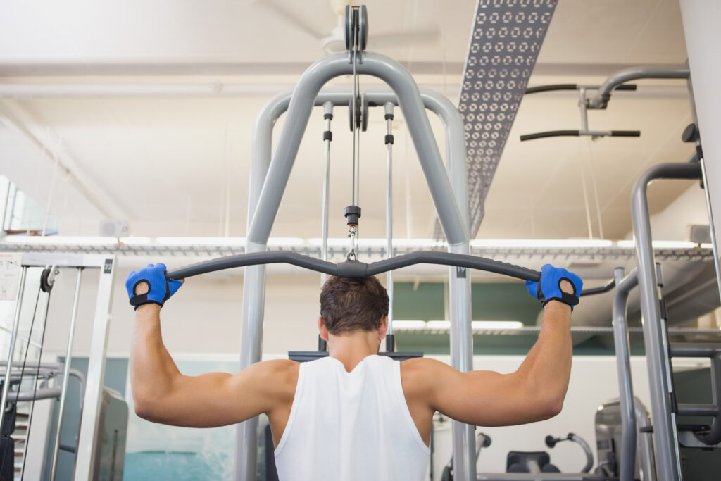 Homem realiza exercício de pulley pronado em máquina de academia, segurando barra ampla atrás da cabeça durante treino de costas.
