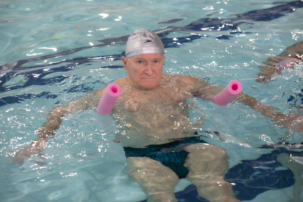 Idoso realiza exercício aquático em piscina com flutuadores de espuma.